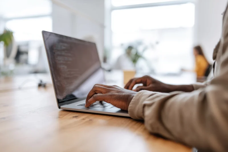 Man typing code on laptop at wooden table in bright office