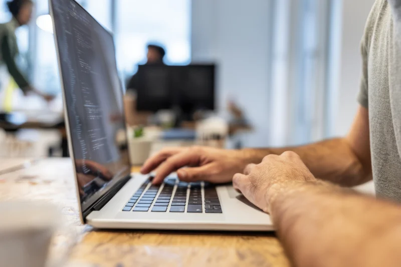 Man typing code on laptop at wooden desk in bright office workspace