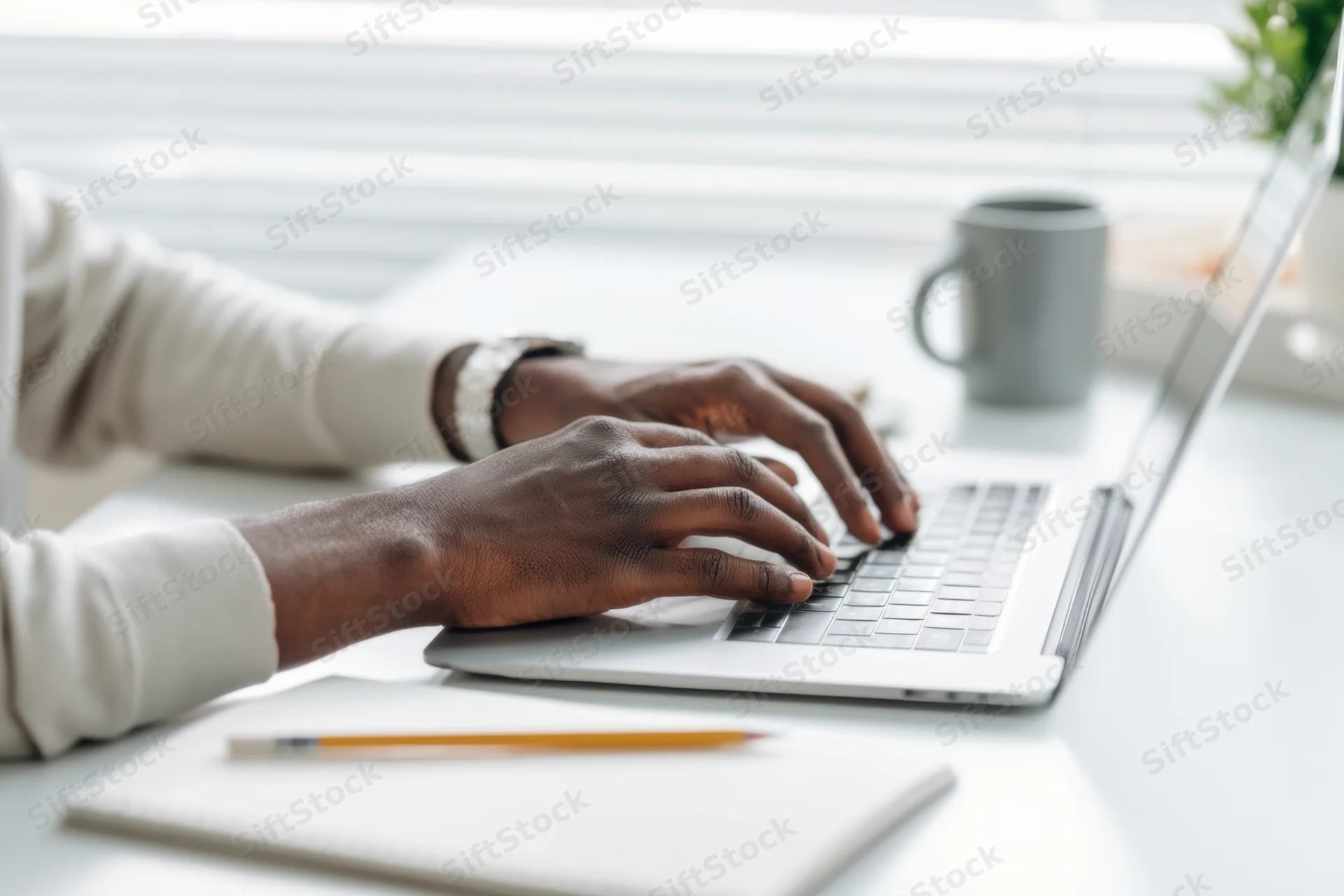 Hands typing on a silver laptop at a bright tidy desk with notebook and mug nearby