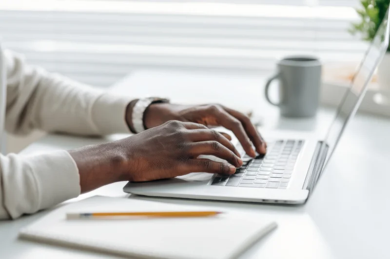 Hands typing on a silver laptop at a bright tidy desk with notebook and mug nearby