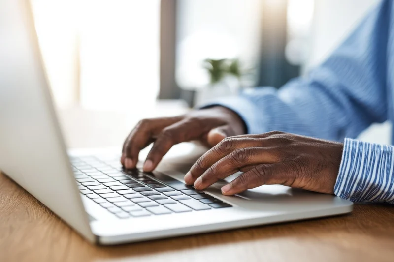 Hands typing on a silver laptop at a wooden desk in an office setting