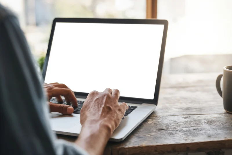 Man typing on silver laptop with blank white screen at wooden desk near window