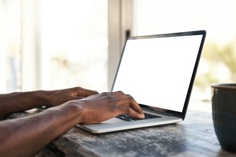 Man typing on silver laptop with blank white screen at wooden desk near window