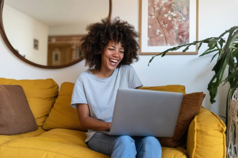 Woman laughing while using laptop on yellow sofa in living room