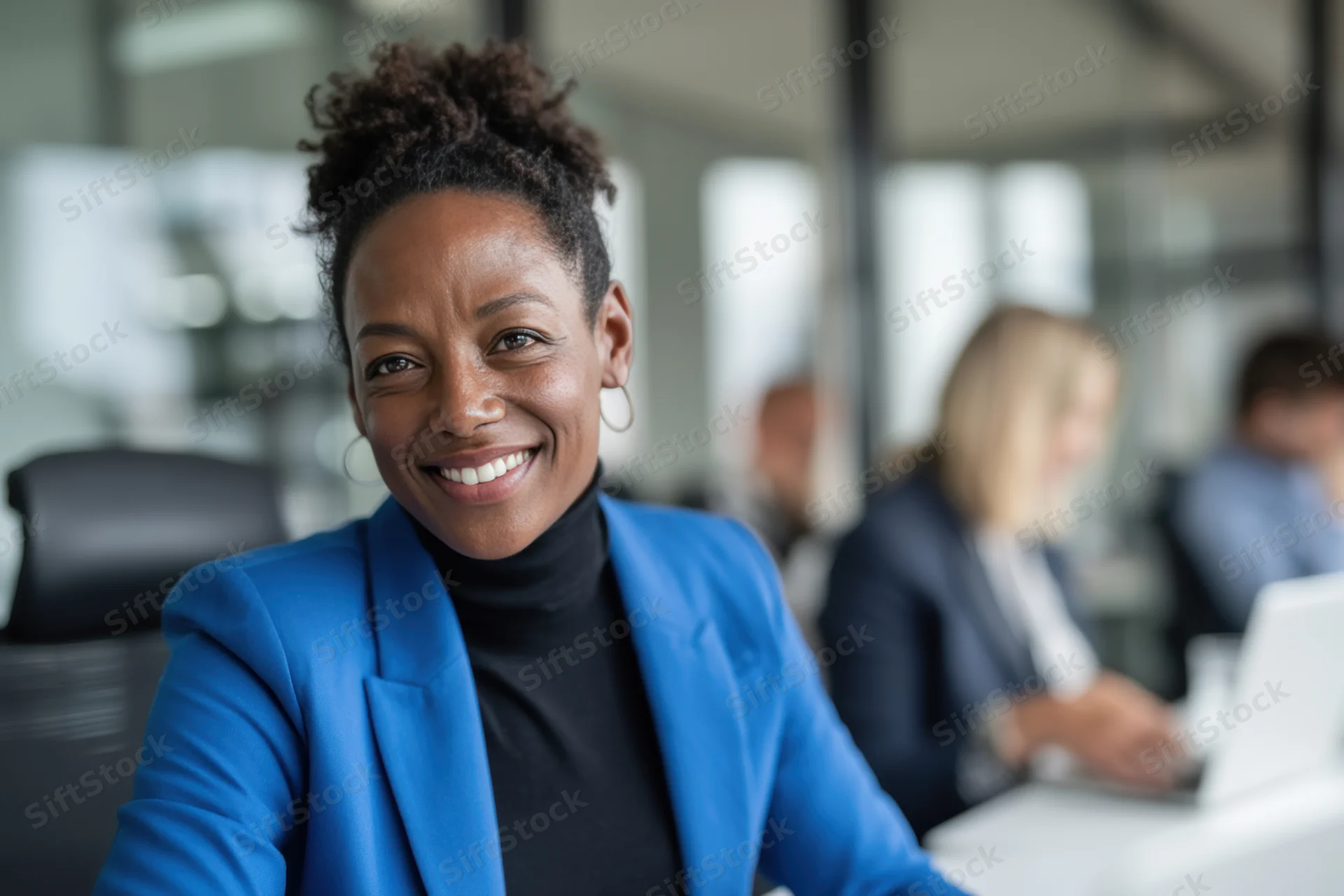 Smiling woman in blue blazer sitting at office desk with colleagues working in background