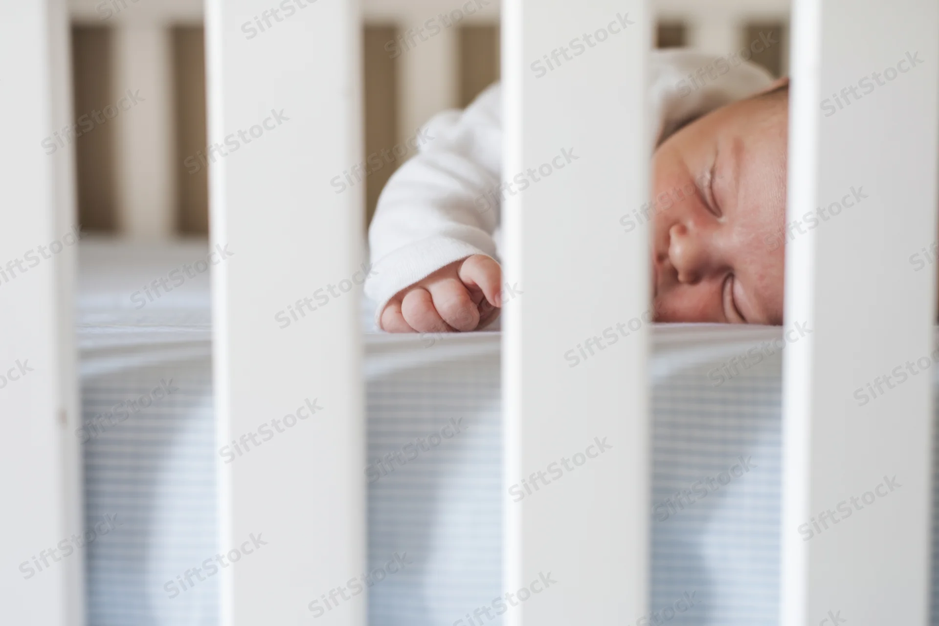Sleeping newborn in white crib with tiny hand reaching through slats