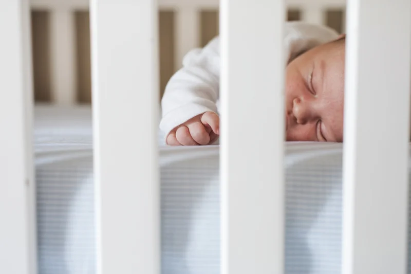 Sleeping newborn in white crib with tiny hand reaching through slats