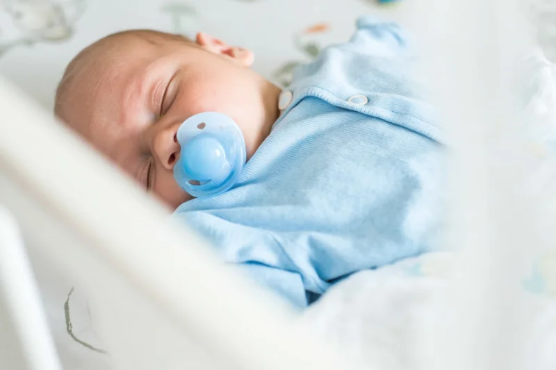 Sleeping newborn in white crib wearing light blue onesie with pacifier