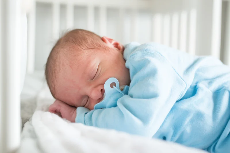 Sleeping newborn baby in white crib wearing light blue onesie with pacifier