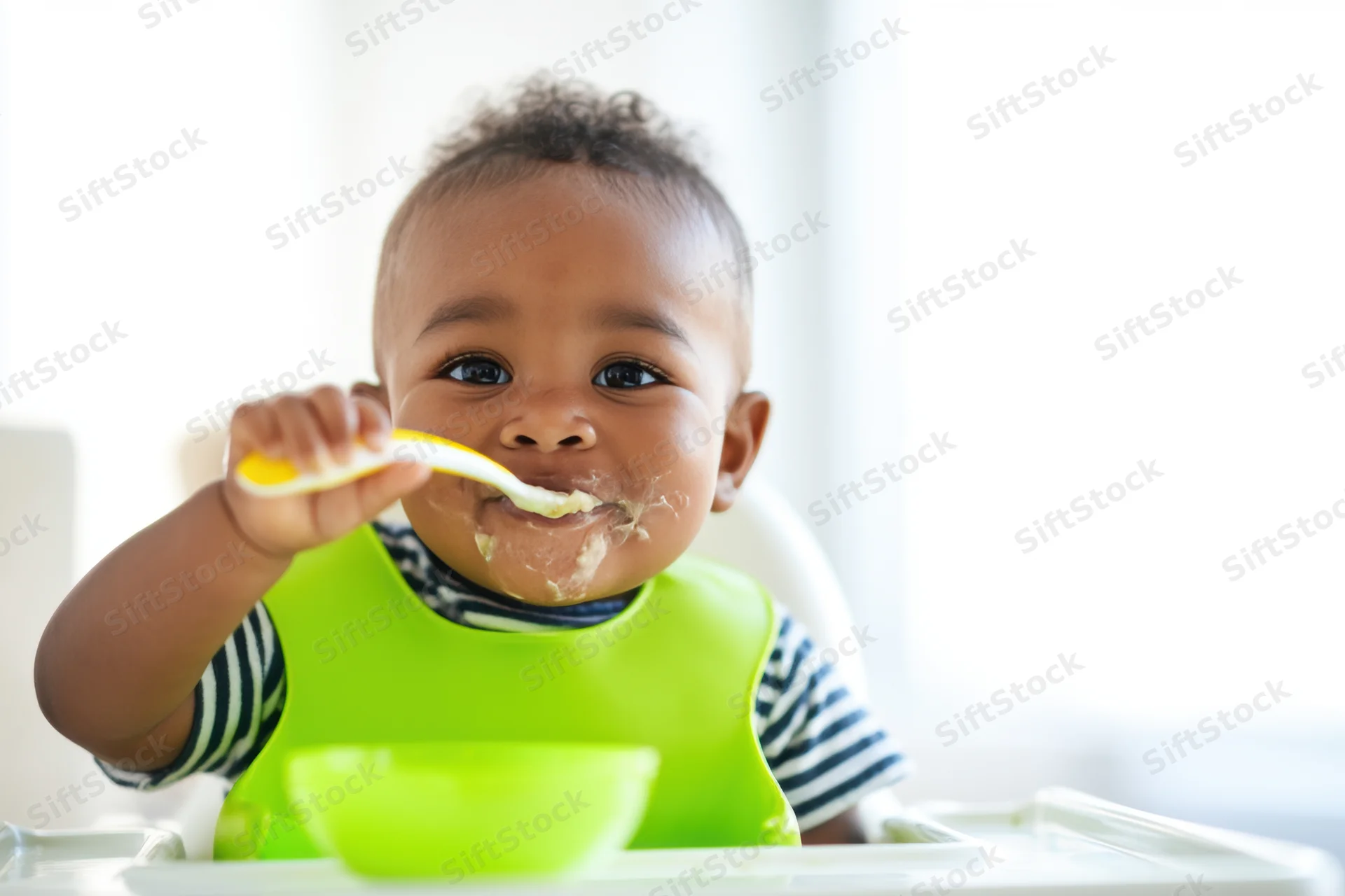 Baby eating puree with spoon in highchair wearing green bib and striped shirt