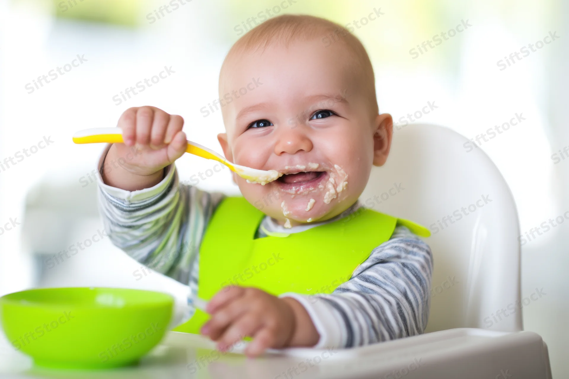 Smiling baby in highchair eating with spoon and green bib