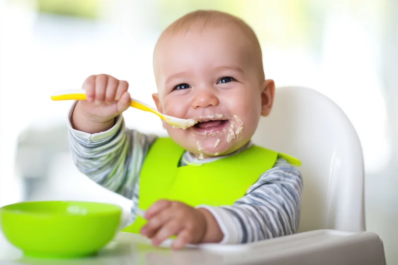 Smiling baby in highchair eating with spoon and green bib