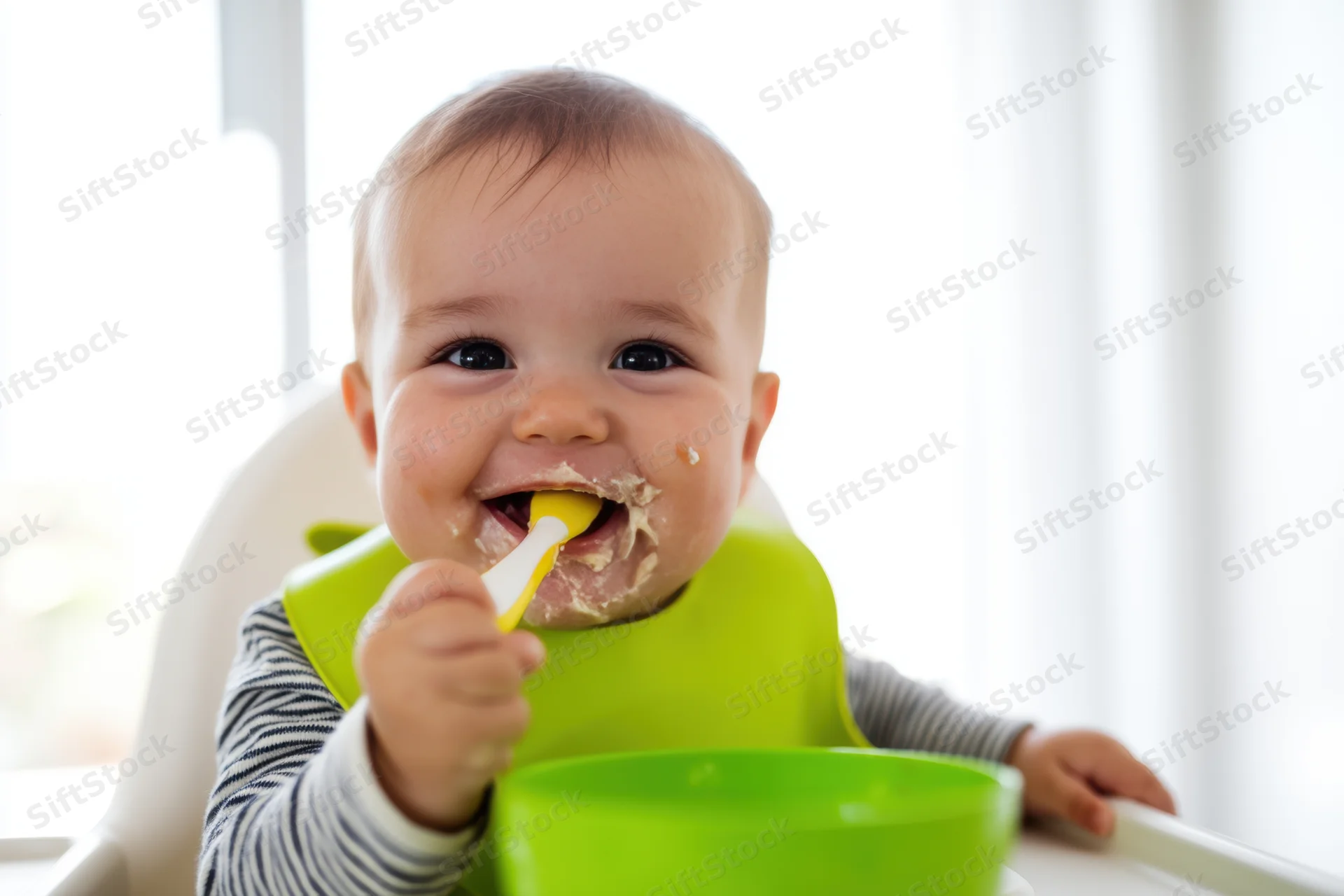 Smiling baby eating mashed food with spoon in highchair wearing green bib and striped shirt