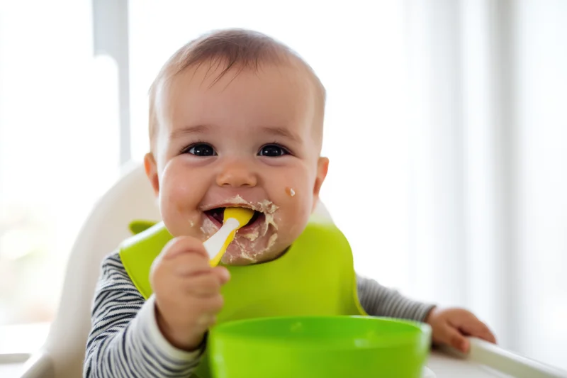 Smiling baby eating mashed food with spoon in highchair wearing green bib and striped shirt