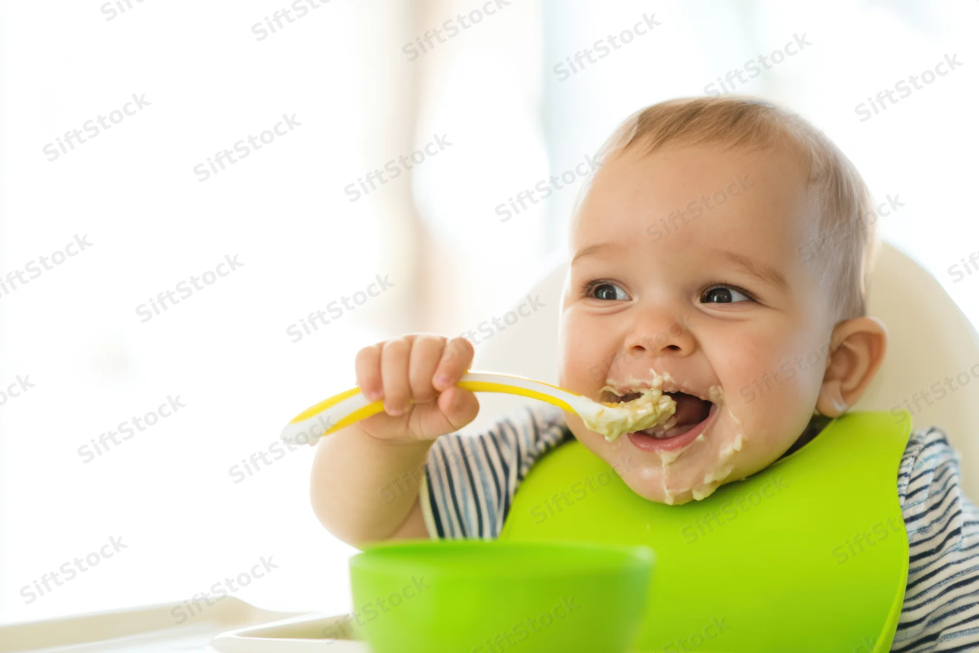Smiling baby in striped shirt eating mashed food with spoon in highchair wearing green bib