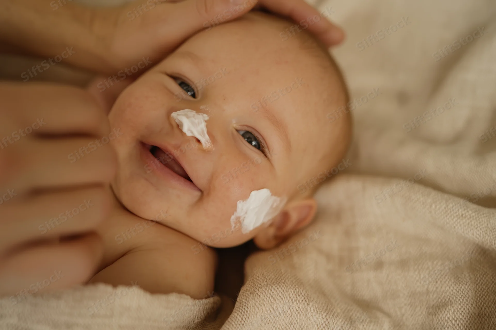 Smiling baby lying on soft beige blanket with white cream on face and hands gently touching head