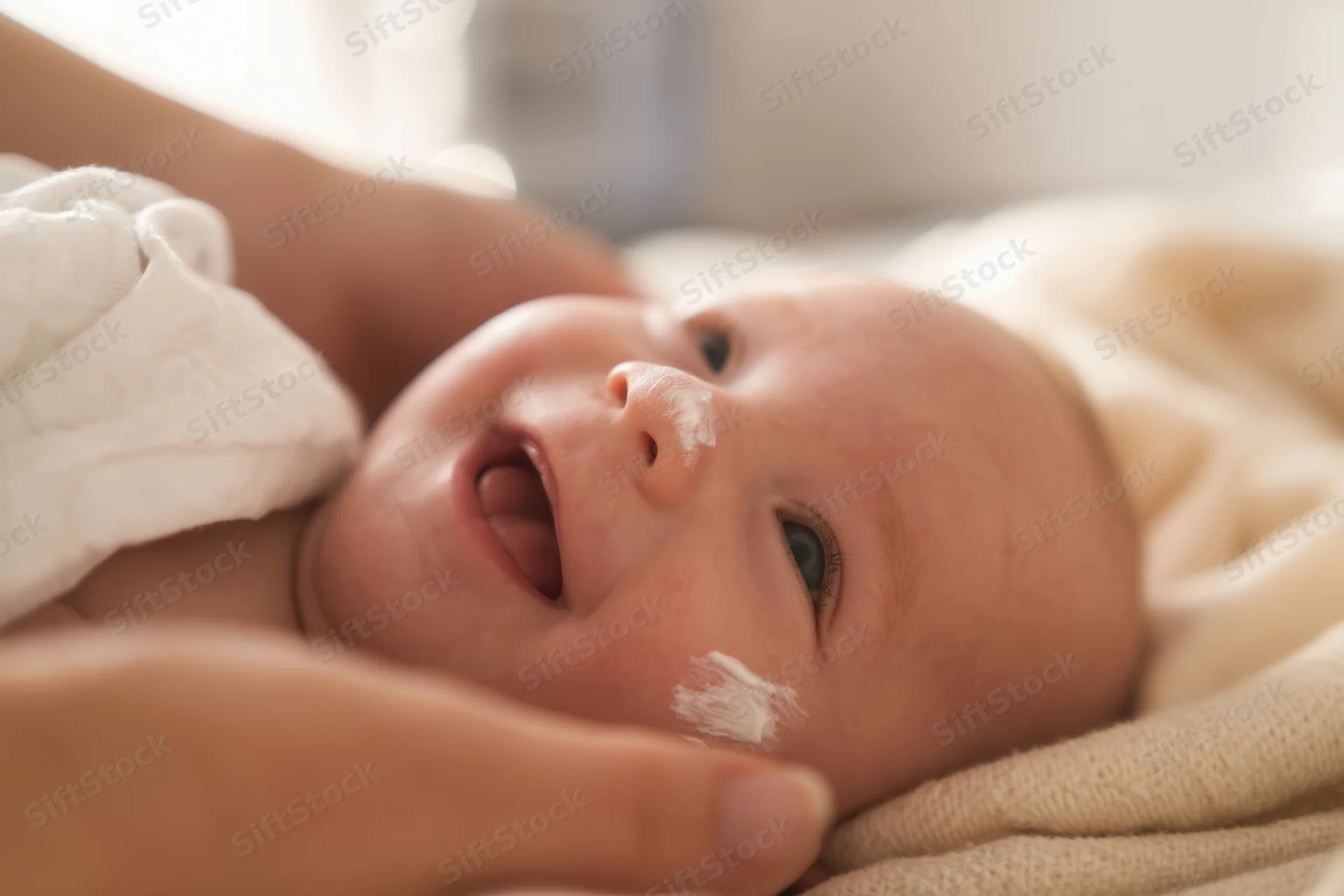 Smiling baby lying on soft beige blanket with white cream on face and hand touching cheek