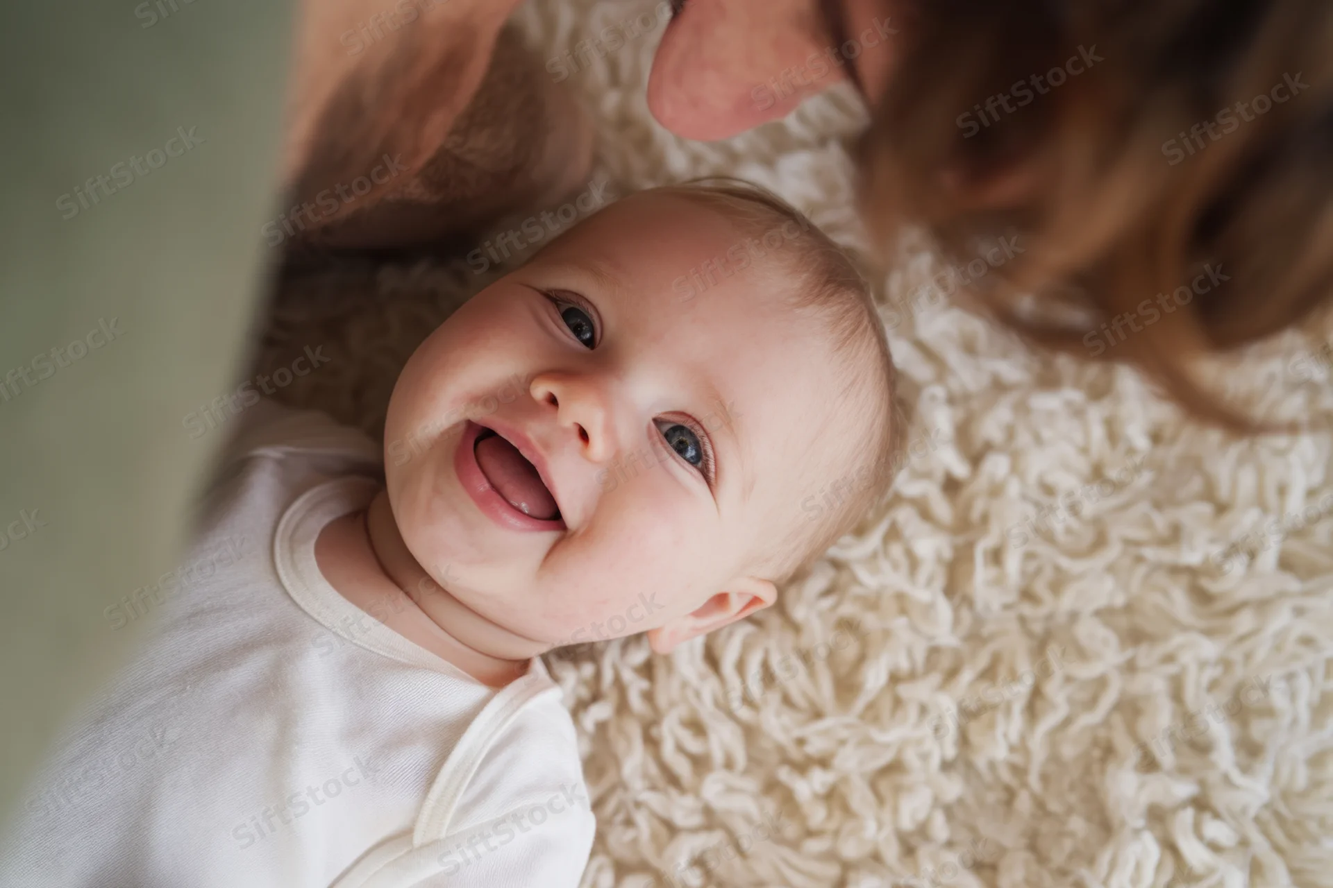 Smiling baby lying on soft shaggy rug with adult leaning over