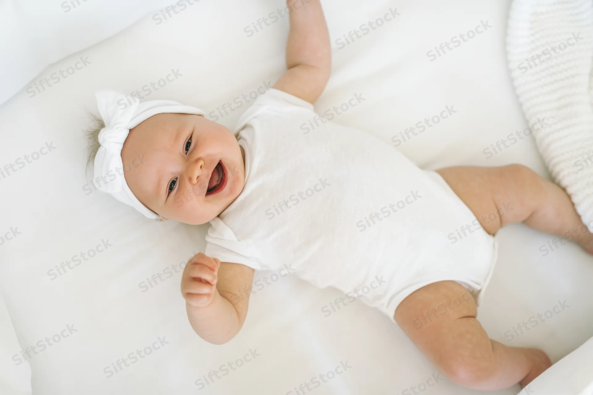 Smiling baby lying on white crib mattress wearing white onesie and headband with bow