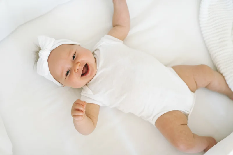 Smiling baby lying on white crib mattress wearing white onesie and headband with bow