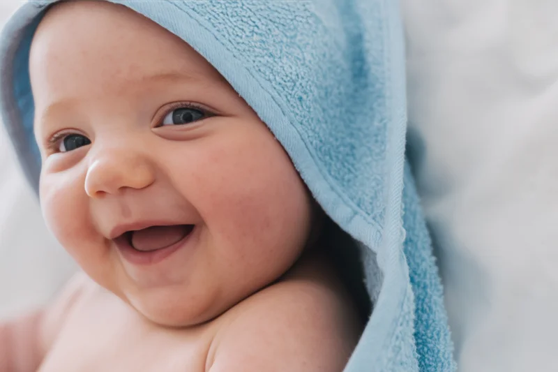 Smiling baby with light blue hooded towel draped over head, close-up portrait indoors