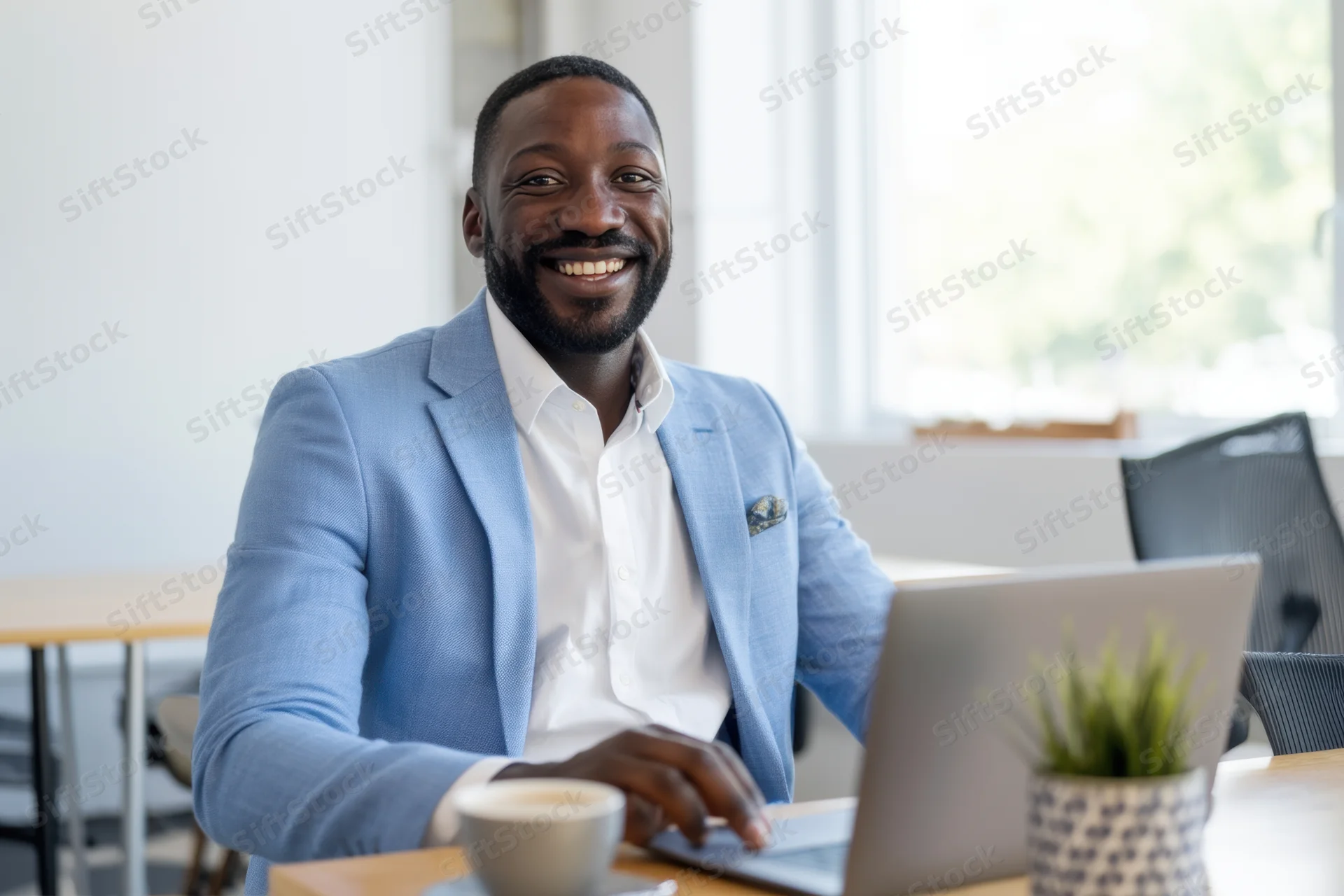 Smiling professional man in light blue blazer working on laptop at office desk with coffee and plant nearby