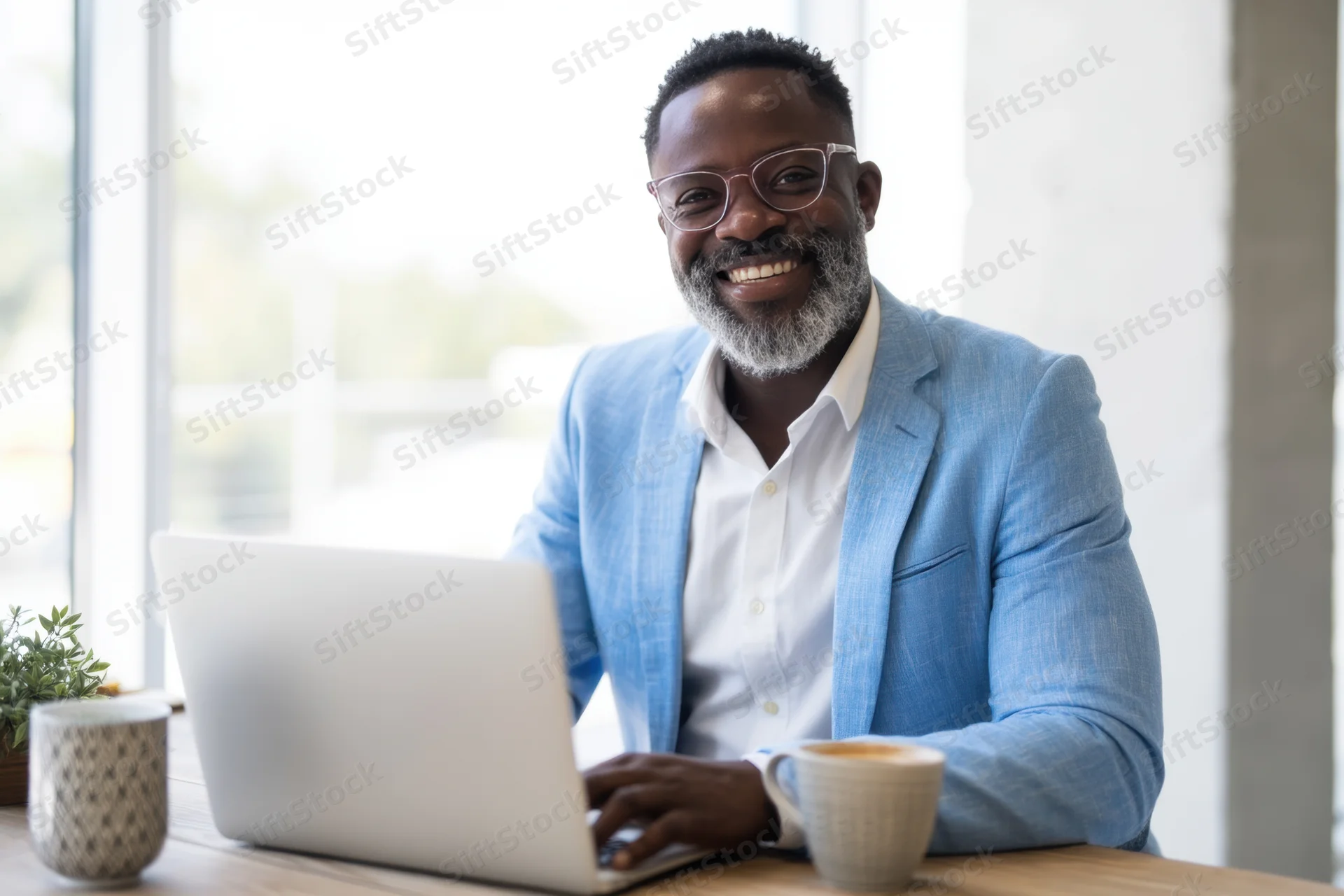 Smiling professional man in light blue blazer working on laptop at desk with coffee mug and plant near window