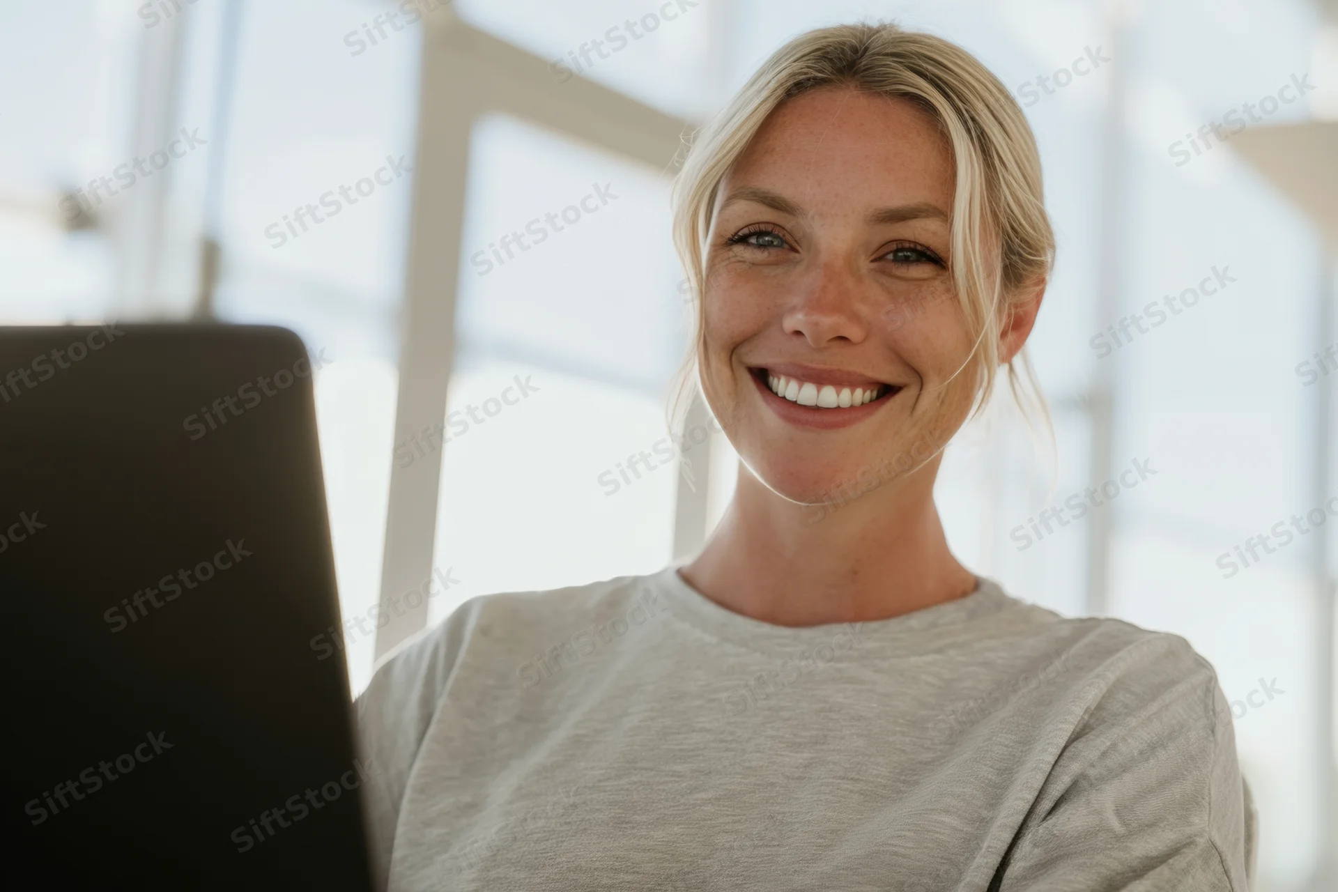 Smiling young woman with blonde hair tied back sitting at laptop in bright office space