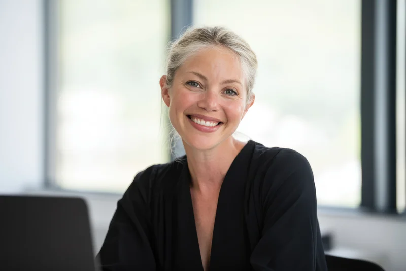 Smiling blonde woman with hair tied back sitting at office desk near window
