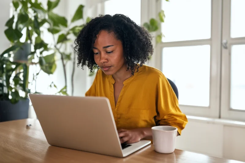 Woman working on laptop at wooden desk in home office with plant and window in background