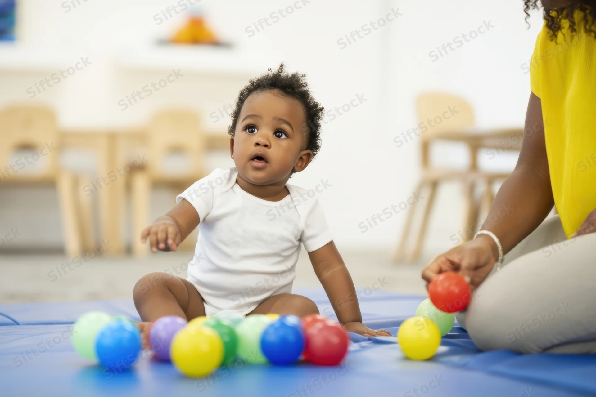Baby sitting on blue padded mat playing with colorful balls while woman interacts nearby