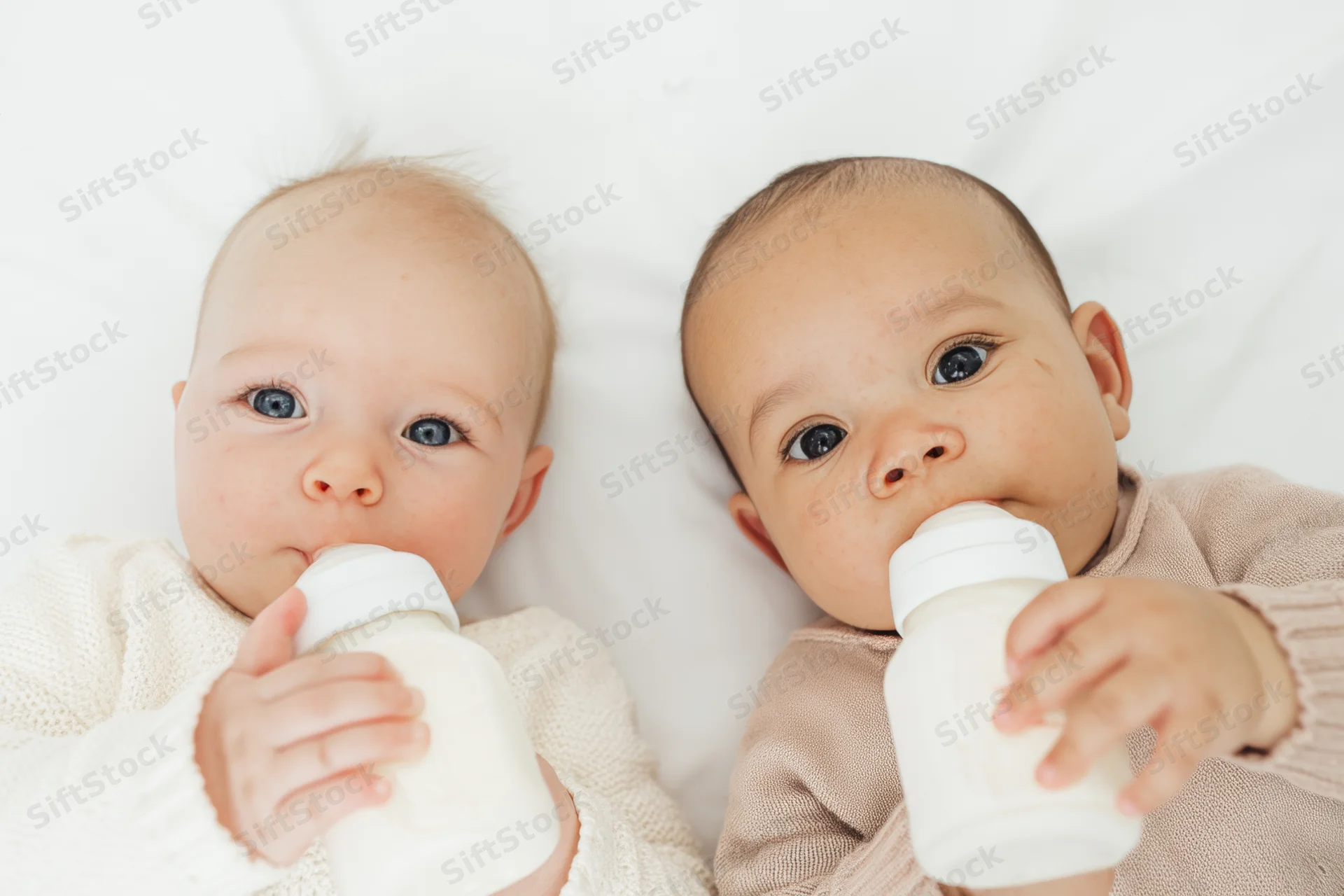 Two babies lying on white surface each drinking from a bottle