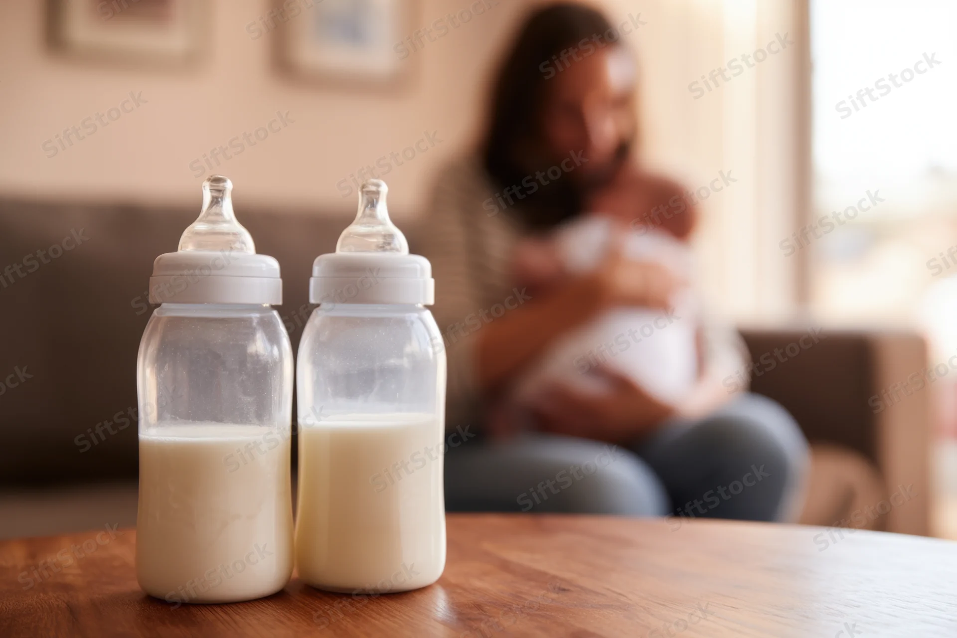 Two baby bottles filled with milk on a wooden table in front of a mother holding a newborn on a sofa