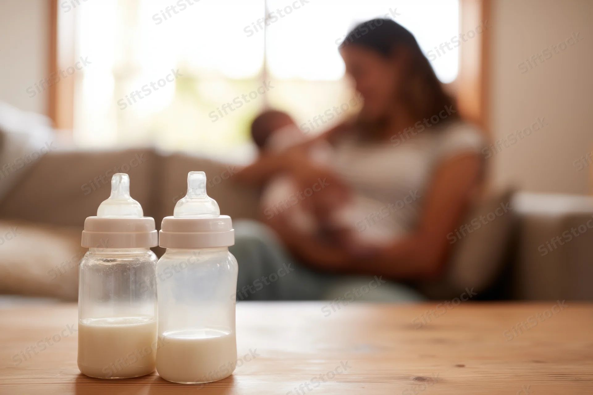 Two baby bottles filled with milk on a wooden table in the foreground with a mother nursing a newborn on a sofa in the background