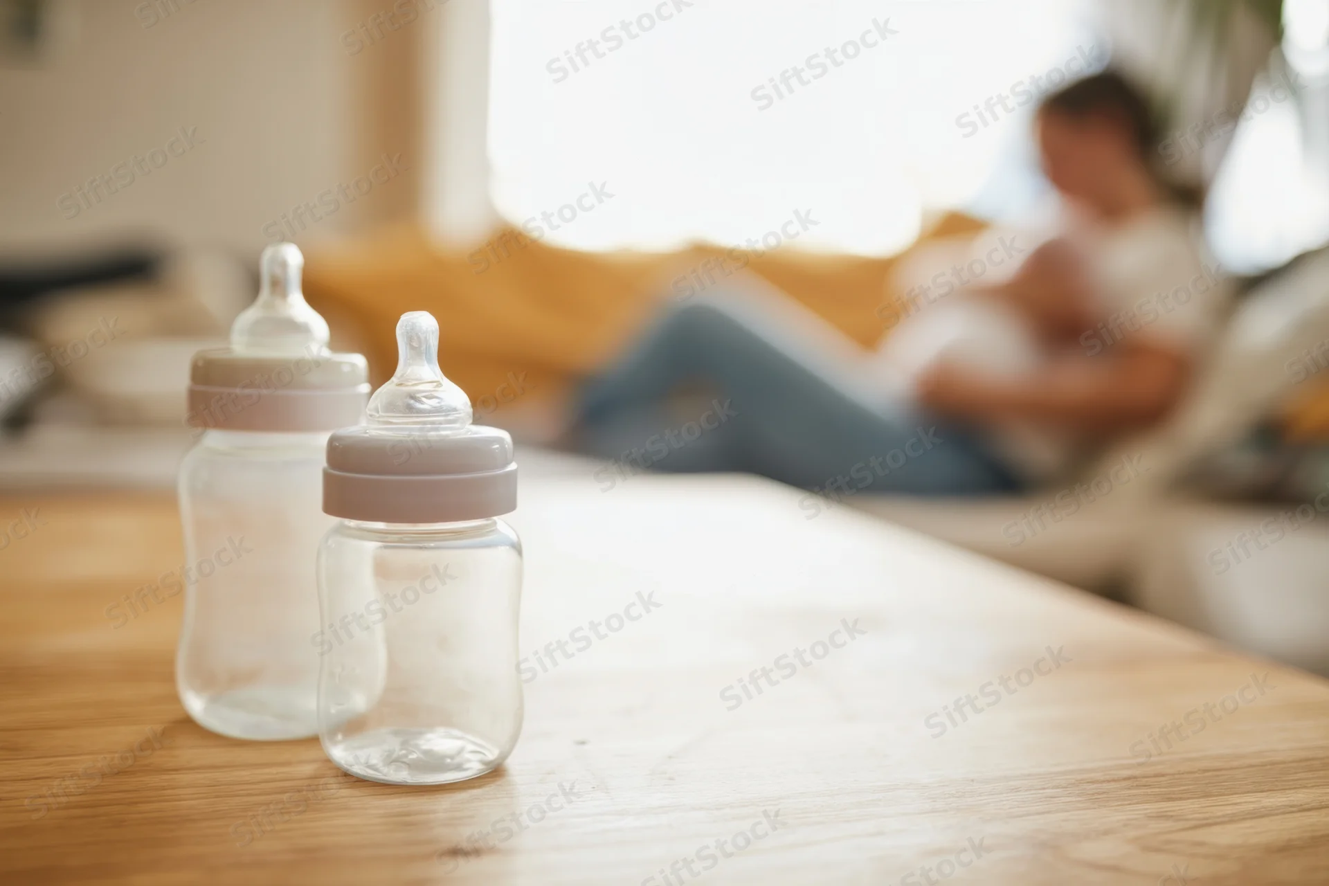 Two baby bottles on a wooden table with a blurred woman breastfeeding in the background