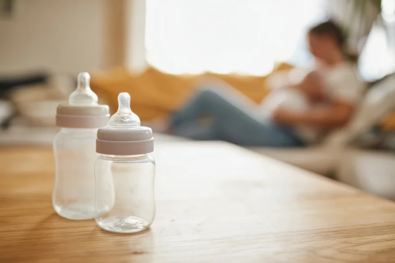 Two baby bottles on a wooden table with a blurred woman breastfeeding in the background