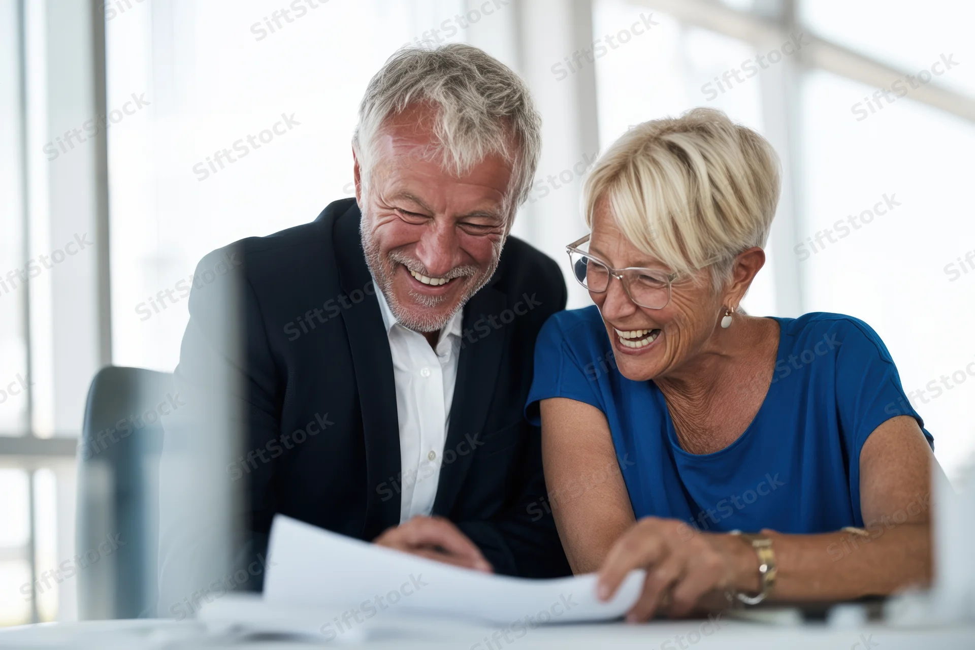 Two senior colleagues laughing while reviewing documents at a bright modern office desk