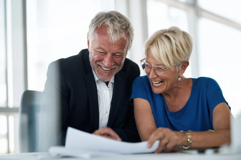 Two senior colleagues laughing while reviewing documents at a bright modern office desk