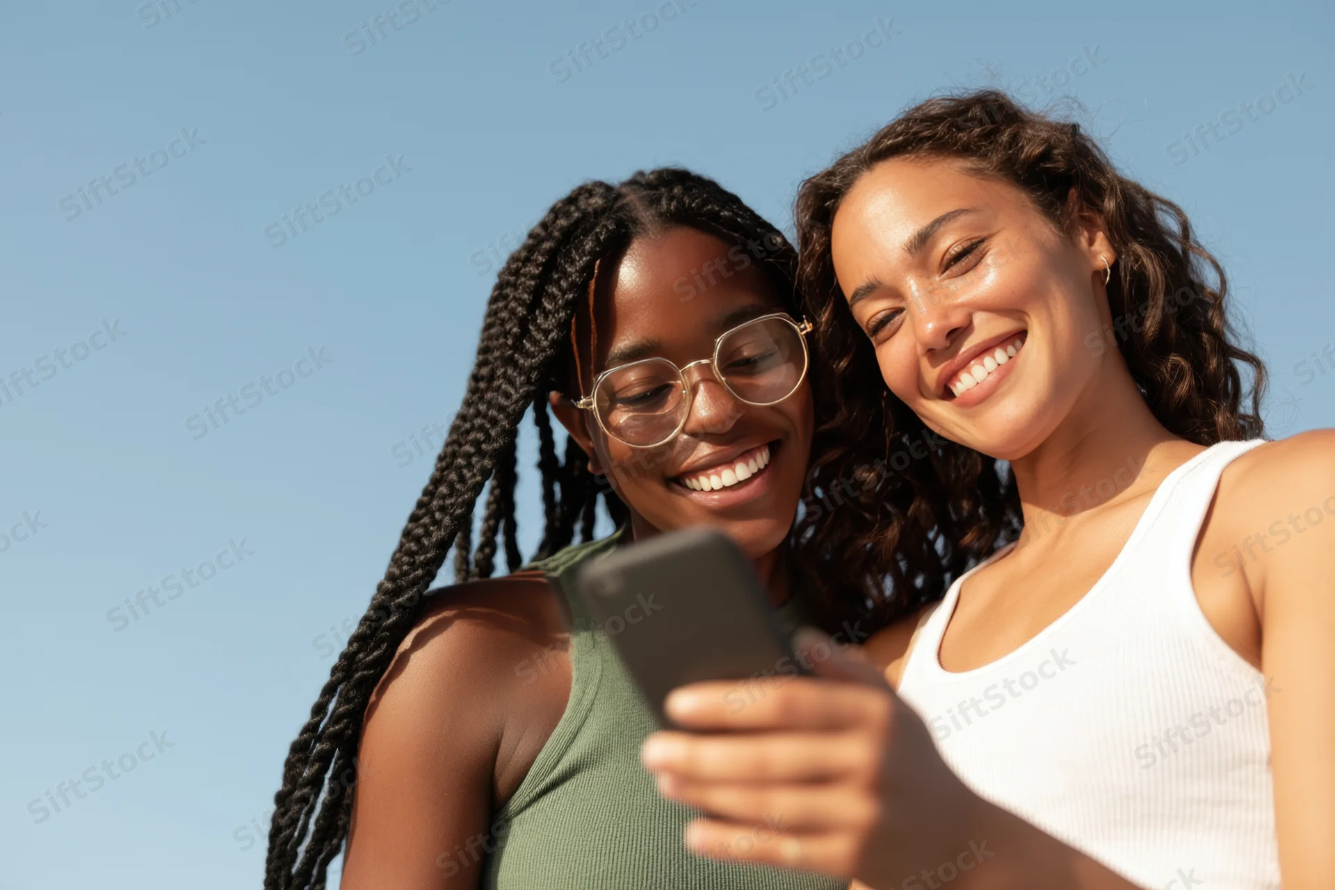 Two women smiling and laughing while looking at a smartphone outdoors under a clear blue sky