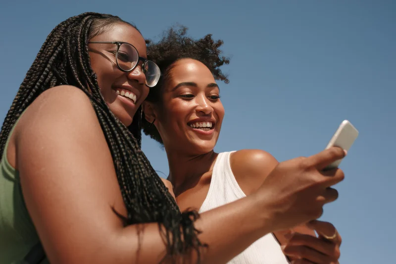 Two women smiling and laughing while looking at a smartphone outdoors under a clear blue sky