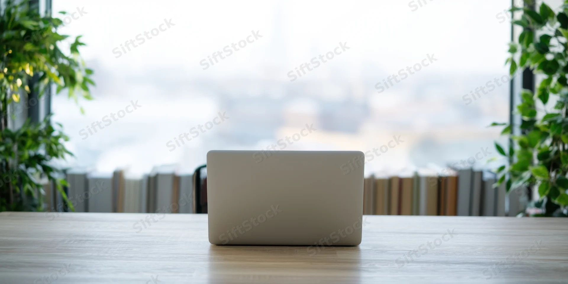 Silver laptop closed on wooden desk in modern office with plants and window view