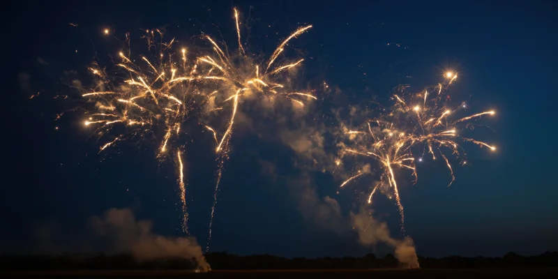 Golden fireworks bursting in the night sky over a dark landscape with smoke trails and sparks
