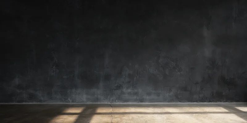 Empty dark studio interior with textured black wall and cracked concrete floor lit by slanted light beams