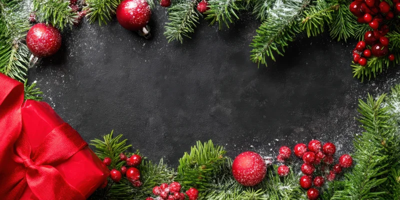 Christmas border of pine branches and red ornaments arranged around dark surface with copyspace