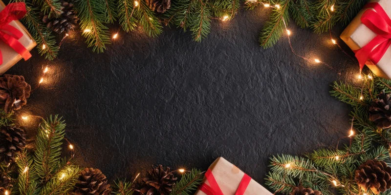 Evergreen branches and pinecones arranged in a festive border with fairy lights and gifts on a dark stone surface