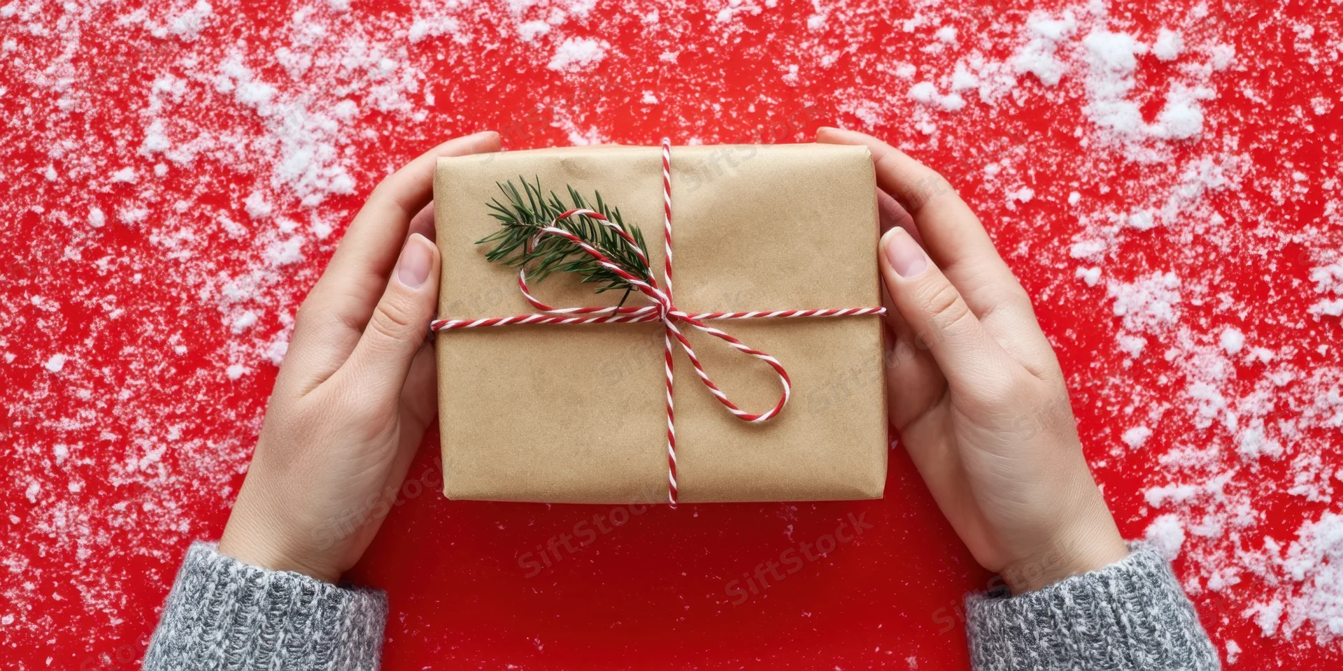 Hands holding a brown gift box wrapped with red and white twine and pine sprig on a red snowy surface