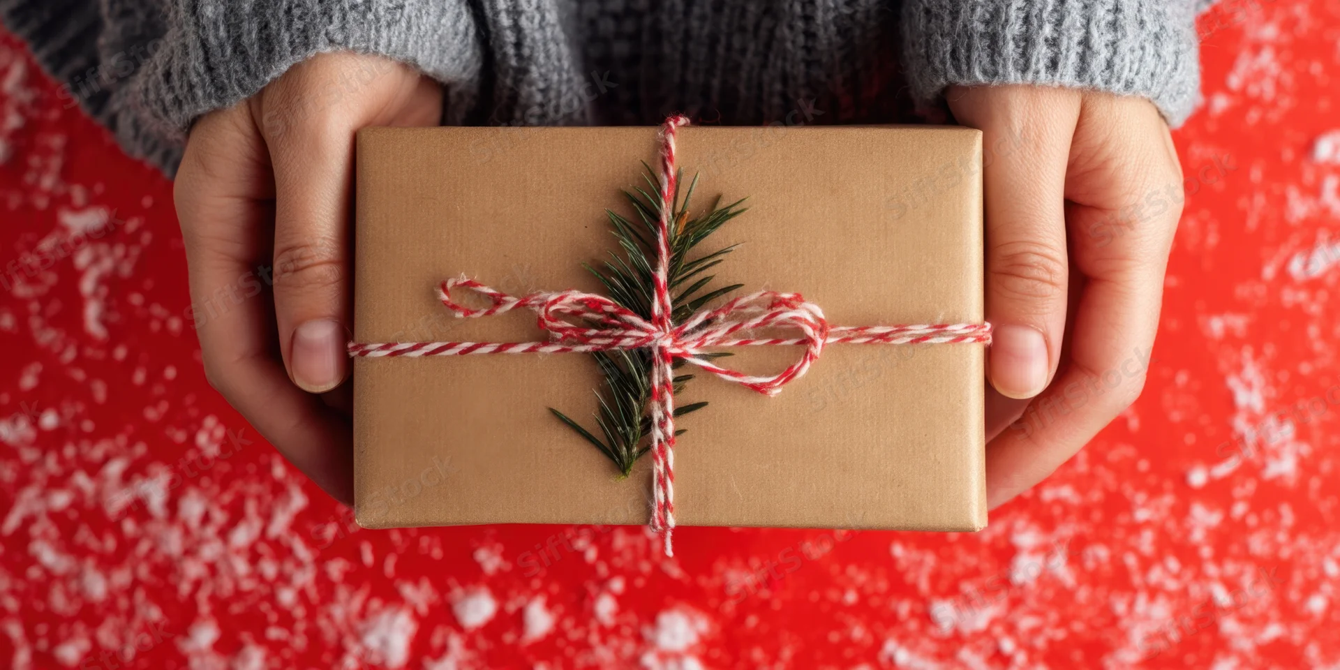 Hands holding a brown gift box wrapped with red and white twine and pine sprig against red snowy background