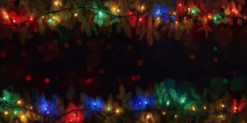 Evergreen branches with colorful string lights arranged as a festive frame against a dark background with bokeh lights and copyspace