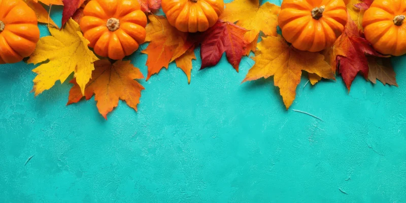 Small orange pumpkins and colorful autumn leaves arranged in a border on a teal surface with copyspace