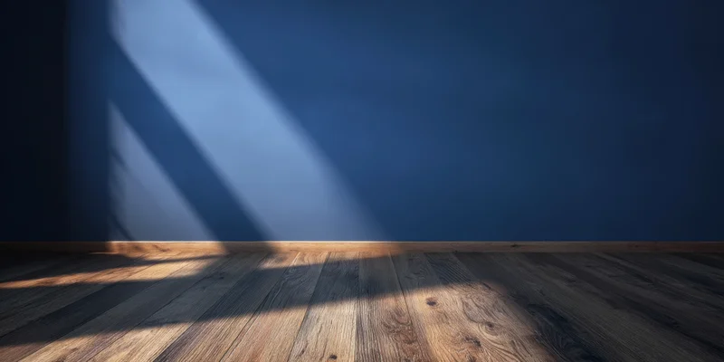 Empty room with deep blue wall and wooden floor lit by diagonal sunlight casting shadows on the floor
